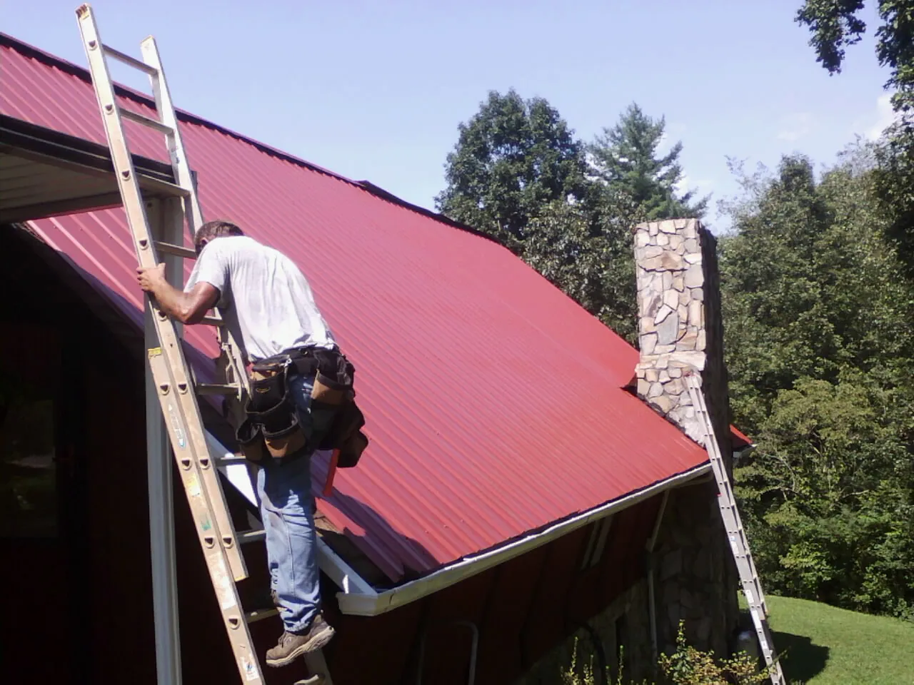 Roofer on a ladder beside a red metal roof and stone chimney — Soffit & Fascia Repair by Roofing Brothers in New Waterford